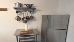 Una cocina compacta con un refrigerador de acero inoxidable, una tabla de cortar de madera sobre una mesa metálica, y ollas y sartenes colgando.