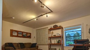 Una sala de estar con piso de madera, un sofá, una mesa de centro, una estantería y un ventanal con vistas a árboles.