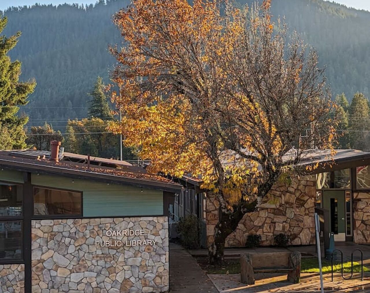 The Oakridge Public Library with stone walls, a wooden deck, and a large tree in the background.