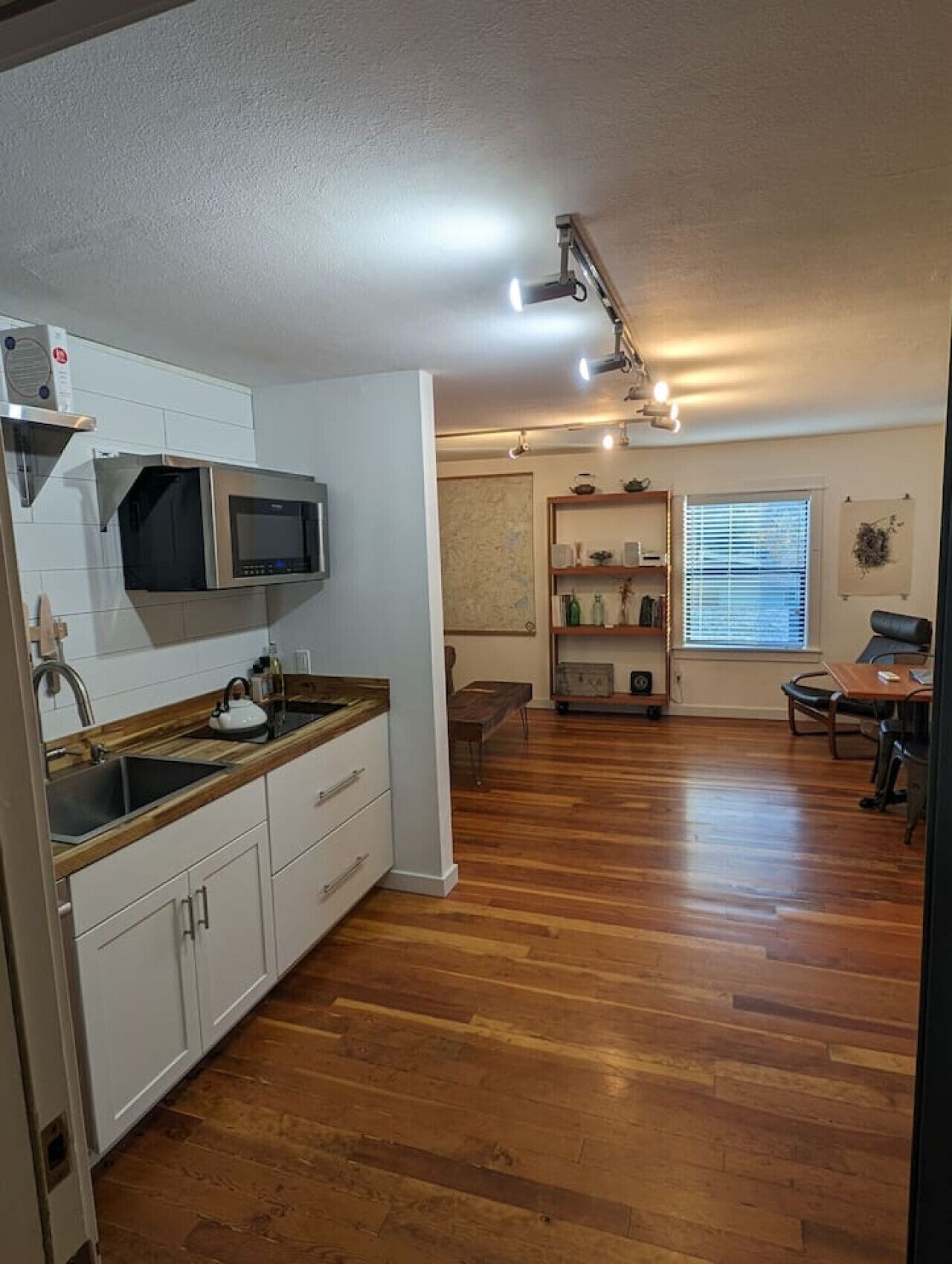 A modern kitchen with white cabinets, a microwave, and a wooden countertop. There is a dining area with a table and chairs, and a window with blinds.