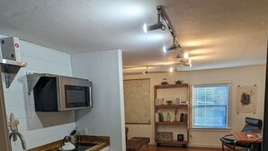 Una cocina moderna con armarios blancos, un microondas y una encimera de madera. Hay un comedor con una mesa y sillas, y una ventana con persianas.