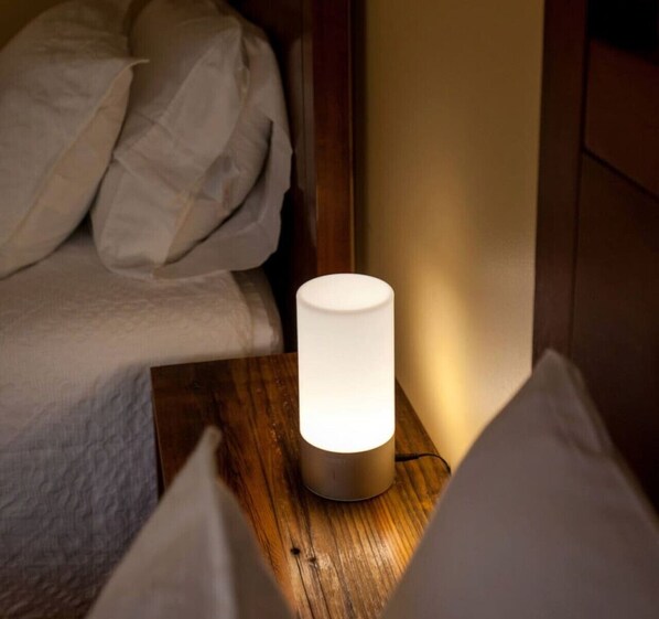 A bedside table with a cylindrical lamp, a bed with white bedding, and a wooden headboard.