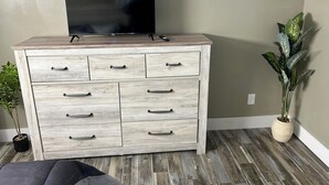 A wooden dresser with drawers and a flat-screen TV on top, a potted plant, and a grey sofa. - Sub Level Effiency (Geneva)