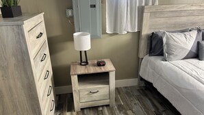 A bedroom with a wooden dresser, a bedside table with a lamp, a clock, and a bed with pillows. - Sub Level Effiency (Geneva)