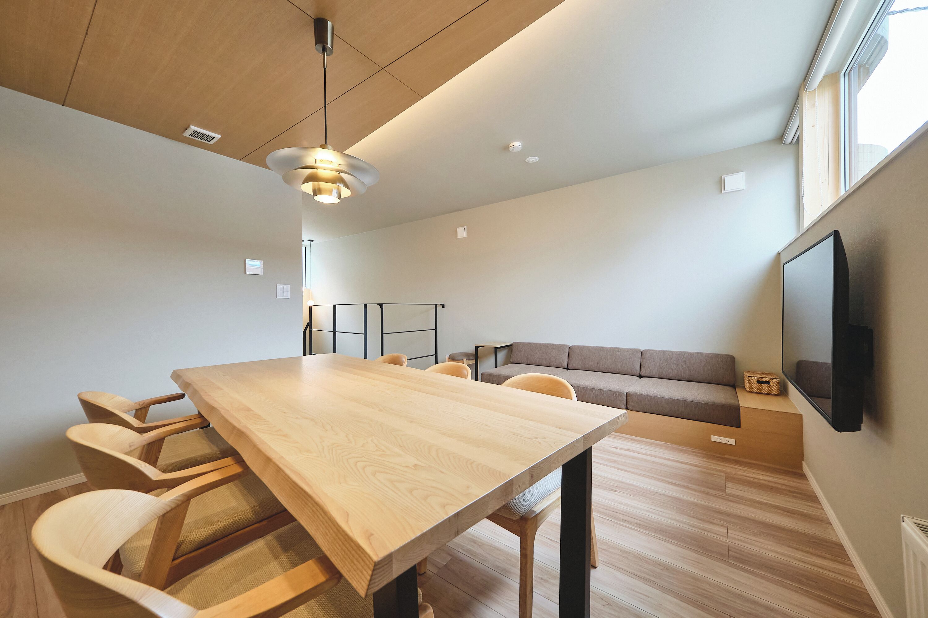 A modern living room with a wooden dining table, cushioned chairs, a sofa, and a mounted TV.