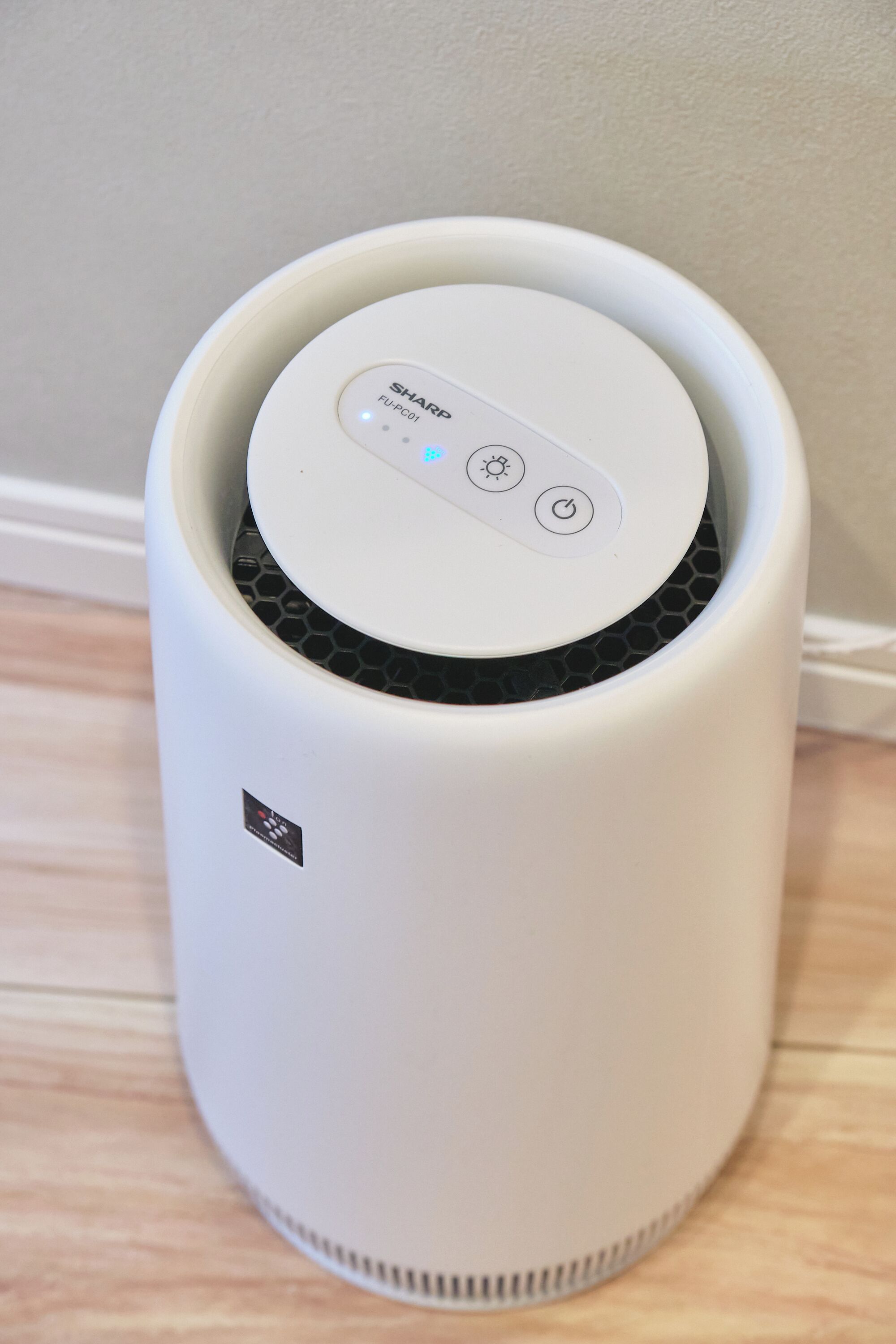 A white air purifier with a control panel on top, placed on a wooden floor against a wall.