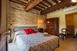 A bedroom with a stone wall, wooden beams, a bed with patterned bedding, a wooden wardrobe, and a bedside table with a lamp. - Villa le Moie by MMega (Arcevia)