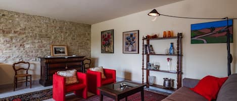 Una sala de estar con pared de piedra, sofás rojo y gris, una mesa de centro de madera y una estantería con diversos objetos.