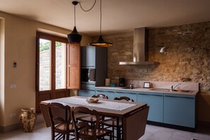 Une cuisine avec un mur en pierre, une table à manger avec des chaises, un luminaire suspendu et un four encastré.