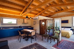 A room with a wooden ceiling, a dining area with chairs, a bed with a blue headboard, and a window.