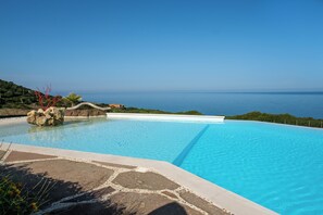 A swimming pool with clear blue water, surrounded by a stone deck and a view of the sea.