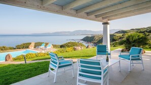 Eine Terrasse im Freien mit weißen Stühlen und einem Tisch, die auf einen Pool und eine schöne Aussicht auf Meer und Hügel blickt.