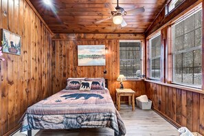 A wooden-paneled room with a bed, a ceiling fan, a window with blinds, and a painting on the wall.
