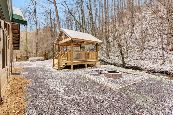 A wooden gazebo with a bench and a fire pit in a forested area.