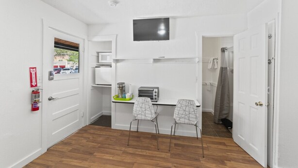 Een kleine keuken met witte kastjes, een magnetron, een broodrooster en een tafeltje met stoelen. Een brandblusser is aan de muur bevestigd.