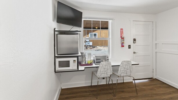 A compact kitchen with a microwave, toaster, and a flat-screen TV mounted on the wall. There are two chairs and a window with a view of the outside. - Atomic #6 I Downtown I Historic I Trailer Parking (Moab)