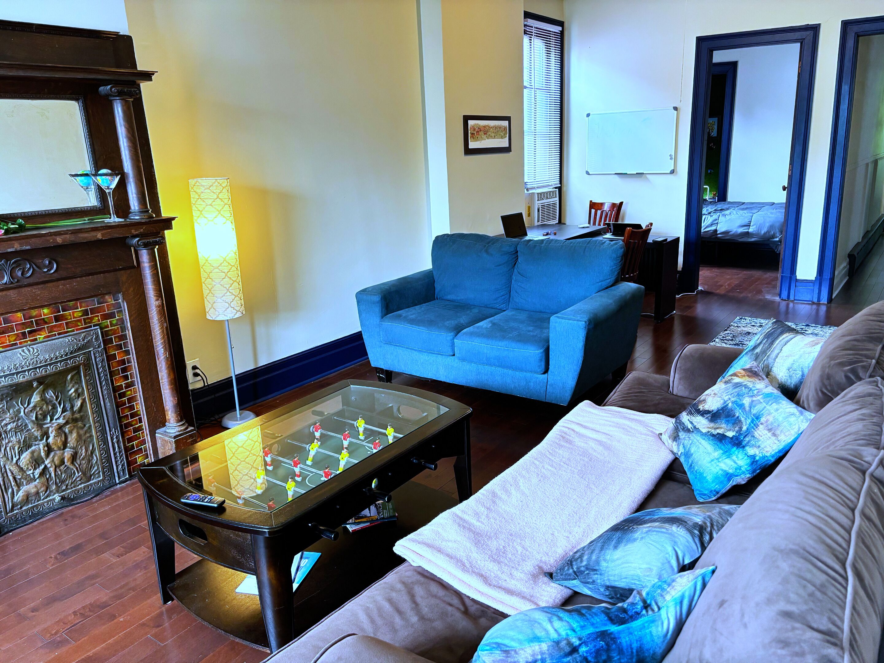 A living room with a blue sofa, a foosball table, a fireplace, and a large window with blinds.