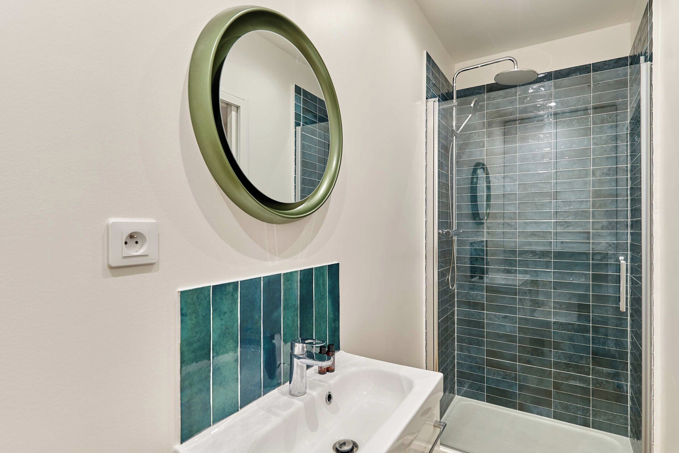 Casa de banho com lavatório branco, espelho com moldura verde, chuveiro com porta de vidro e azulejos verde-água na parede.