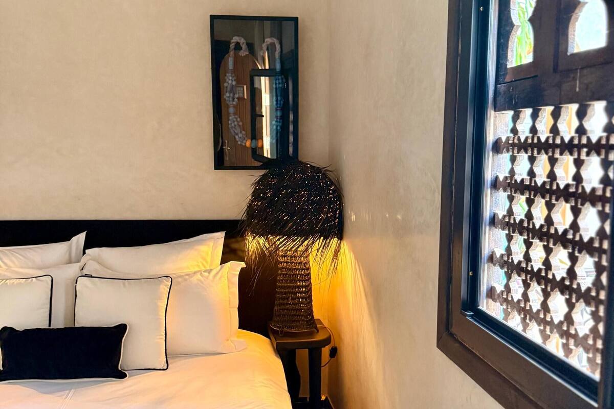 A neatly made bed with white and dark blue bedding, a small table with a lamp, and a window with a decorative screen.