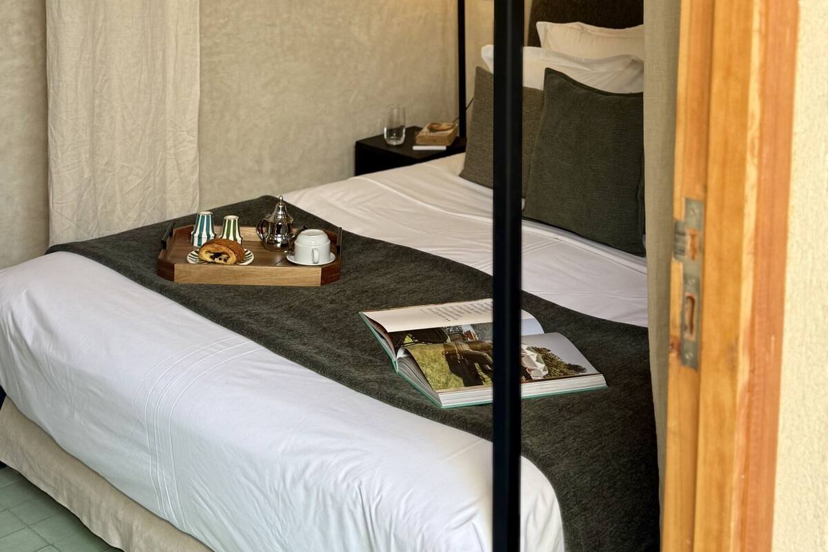 A hotel room with a bed, a bedside table with a lamp, a tray with breakfast items, and a magazine on the bed.