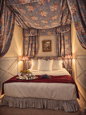 Un lit doté d’un baldaquin à motifs floraux, de draps blancs et d’une table de chevet avec une lampe.