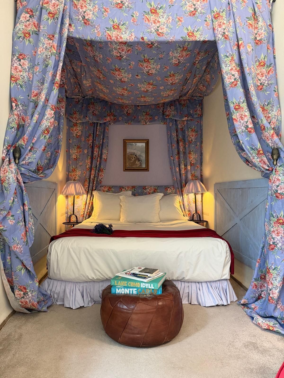 A bedroom with a bed, floral curtains, bedside lamps, a framed picture on the wall, and a stack of books on an ottoman.