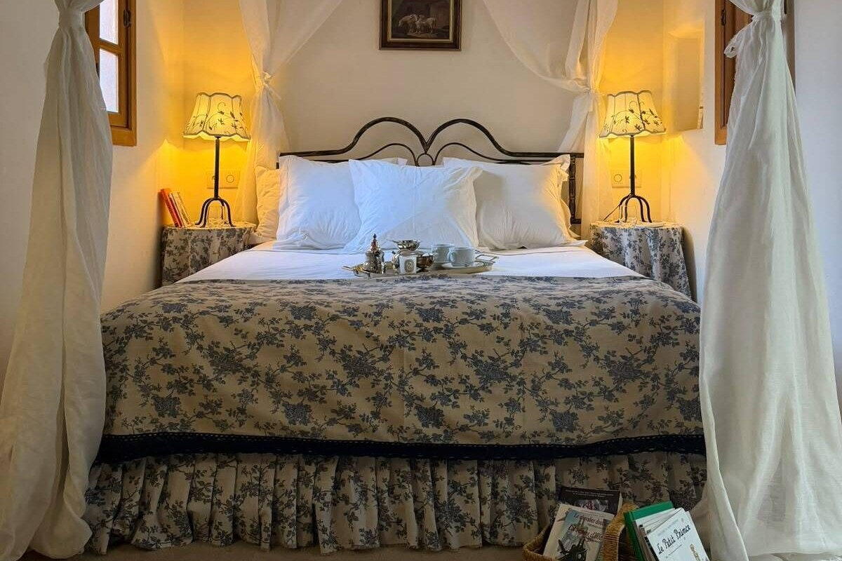 A bed with a canopy, bedside lamps, a tray with tea cups and a teapot, and a basket with books.
