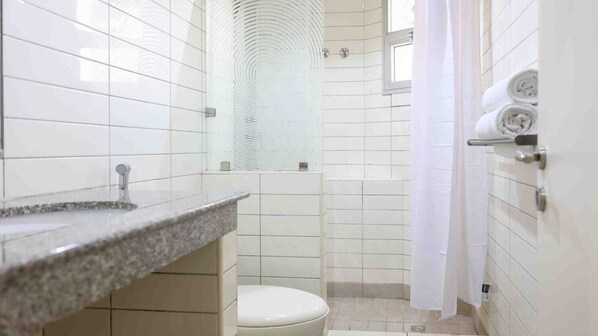 A bathroom with white tiles, a granite countertop, a toilet, a shower with a glass door, and a window. - Code Housing MH8 (Salmiya)