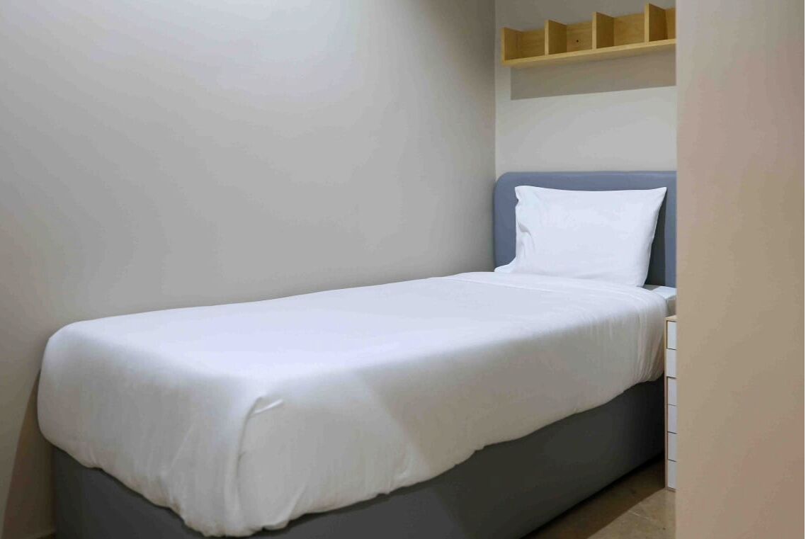 Una habitación pequeña con una cama individual, colchón gris y ropa de cama blanca. Hay una estantería de madera sobre la cama.