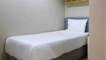 Una habitación pequeña con una cama individual, colchón gris y ropa de cama blanca. Hay una estantería de madera sobre la cama.