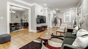 A spacious living room with hardwood floors, a green sofa, a coffee table, and a TV. - Hilltop Retreat w/ Pool, Games & Sunroom (Woodlawn)
