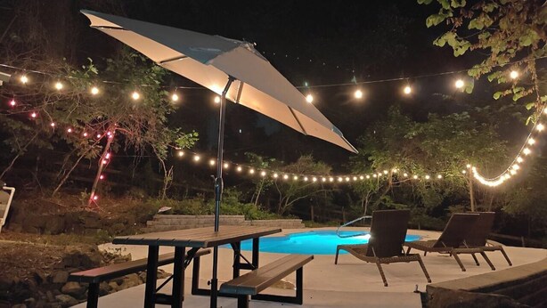 An outdoor pool area with a table and chairs, string lights, and an umbrella. - Hilltop Retreat w/ Pool, Games & Sunroom (Woodlawn)