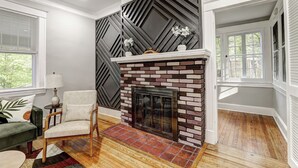 A living room with a fireplace, a green sofa, a white armchair, and a wooden floor. - Hilltop Retreat w/ Pool, Games & Sunroom (Woodlawn)