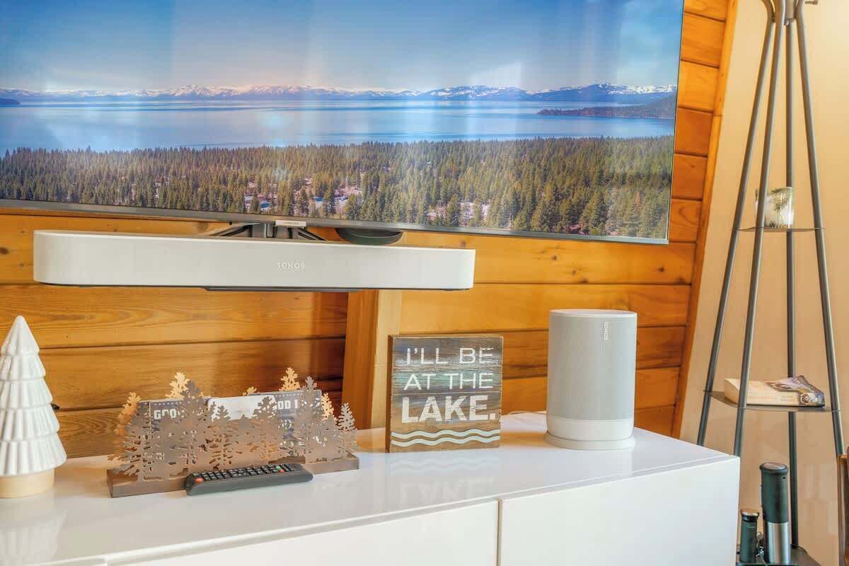 A room with a large TV mounted on a wooden wall, a white entertainment unit with a remote control, a decorative snow scene, and a sign that reads "I'll be at the lake."