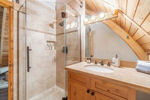A bathroom with a glass-enclosed shower, a wooden vanity with a sink, and a large mirror with lights. - The Canoe Cabin (Incline Village)