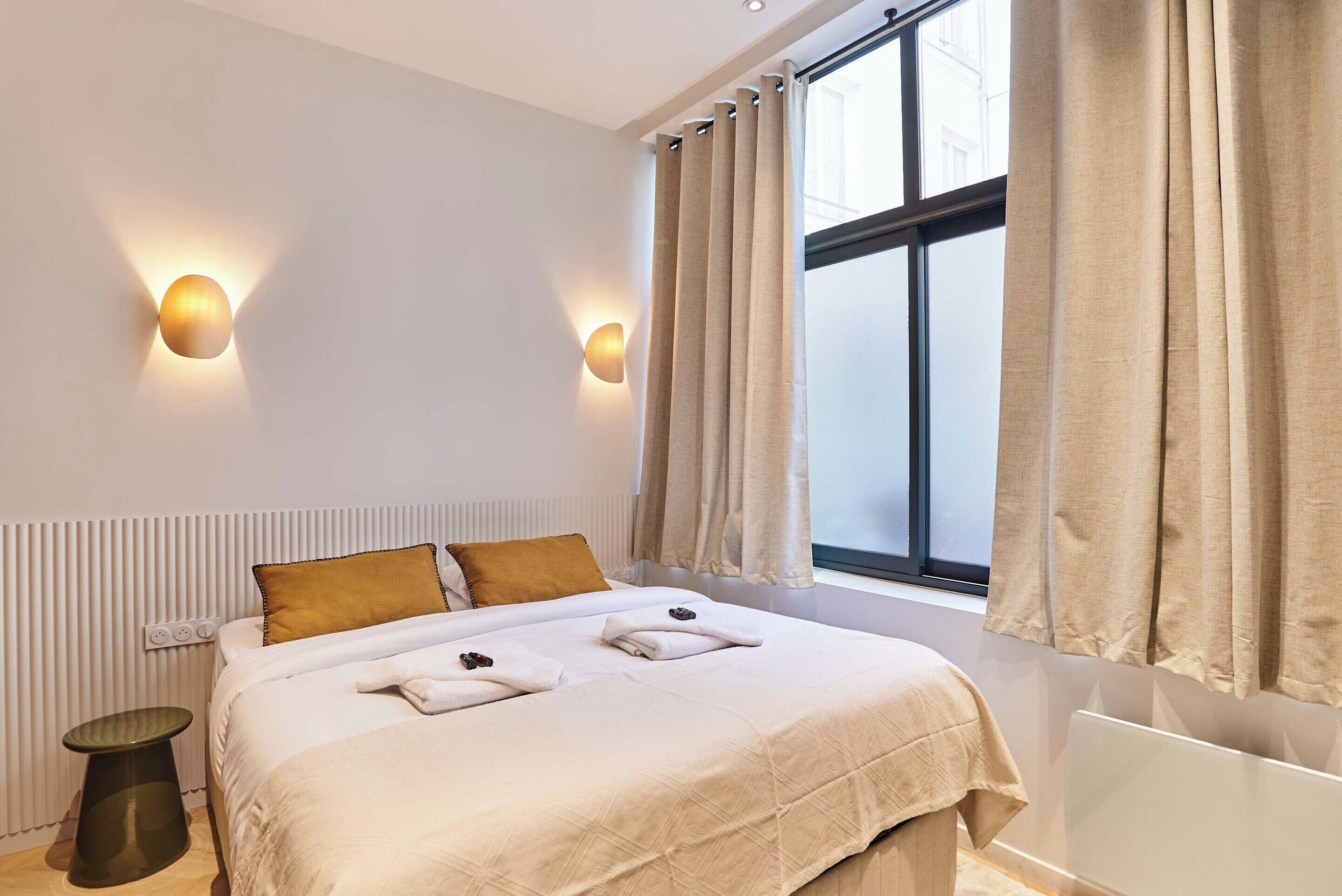 A modern hotel room with a bed, bedside lamps, a window with curtains, and a radiator.