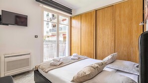 A hotel room with a bed, a TV mounted on the wall, and a window with a view of a balcony. - Modern Apartment - 2br-4p- Croisette Plage (Cannes)