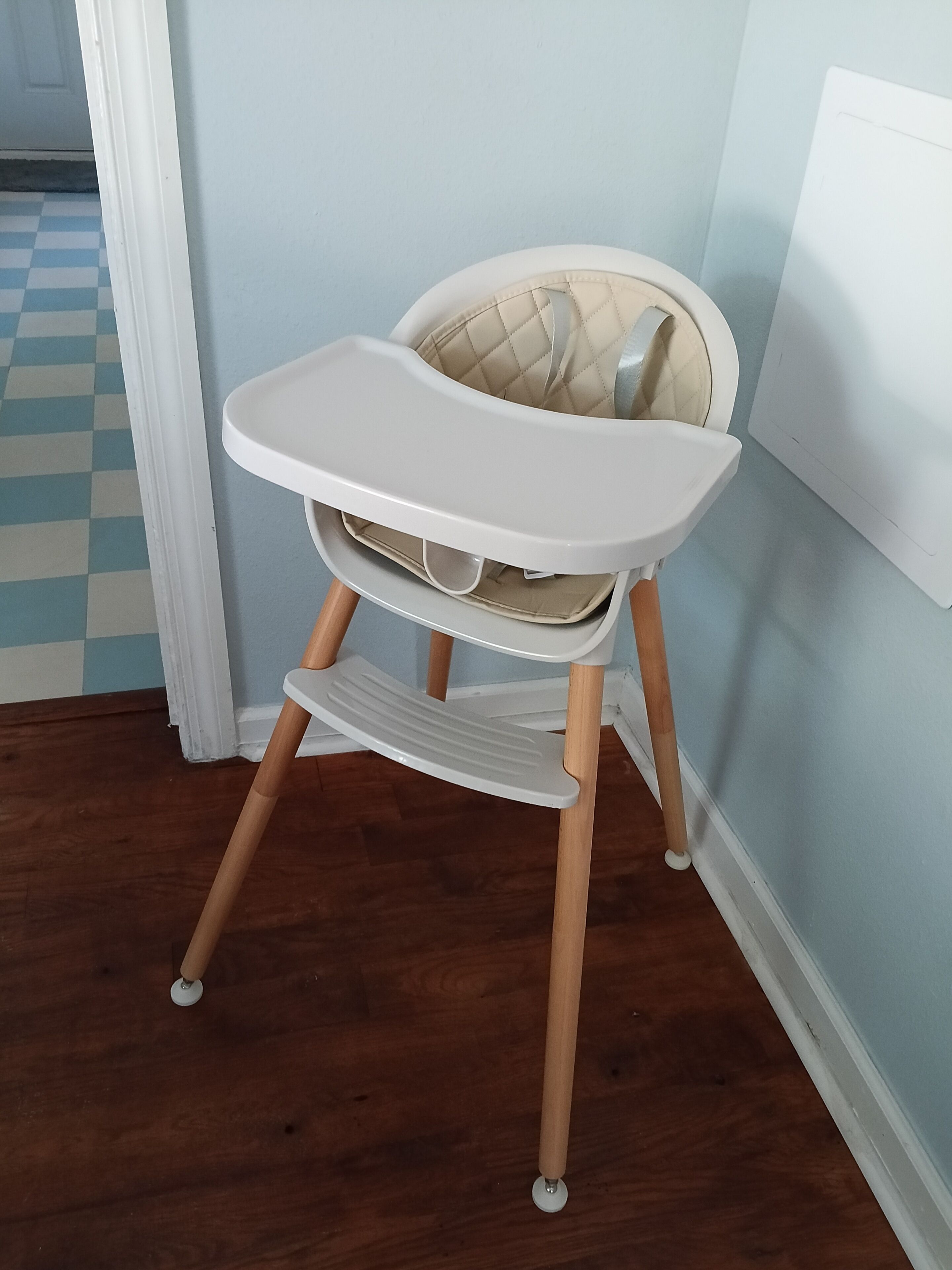 A high chair with a tray and a cushioned seat, positioned against a wall with a checkered floor pattern.