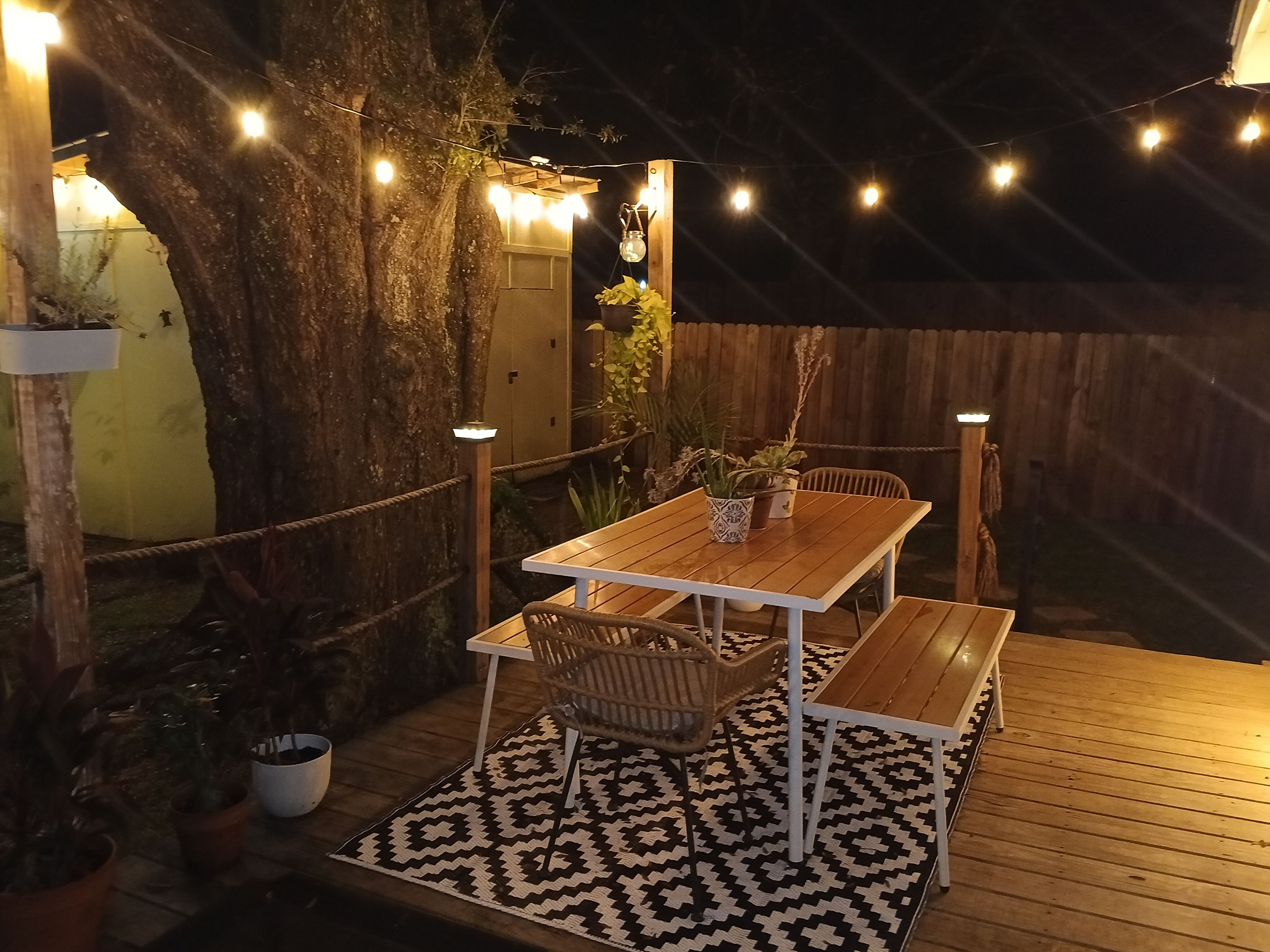 Una terrazza esterna in legno con tavolo e sedie, circondata da piante in vaso e luci decorative.