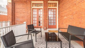 A brick balcony with two double doors, a white railing, and a seating area with two chairs and a table.