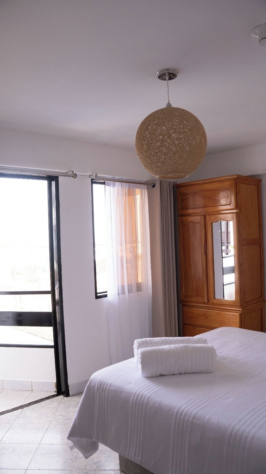 A hotel room with a bed, a wooden wardrobe, a large window with curtains, and a woven ceiling light fixture.