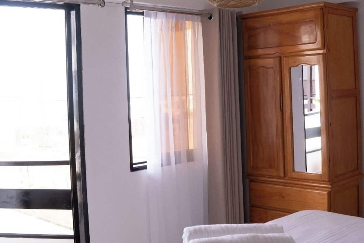 A hotel room with a bed, a wooden wardrobe, a large window with curtains, and a woven ceiling light fixture.