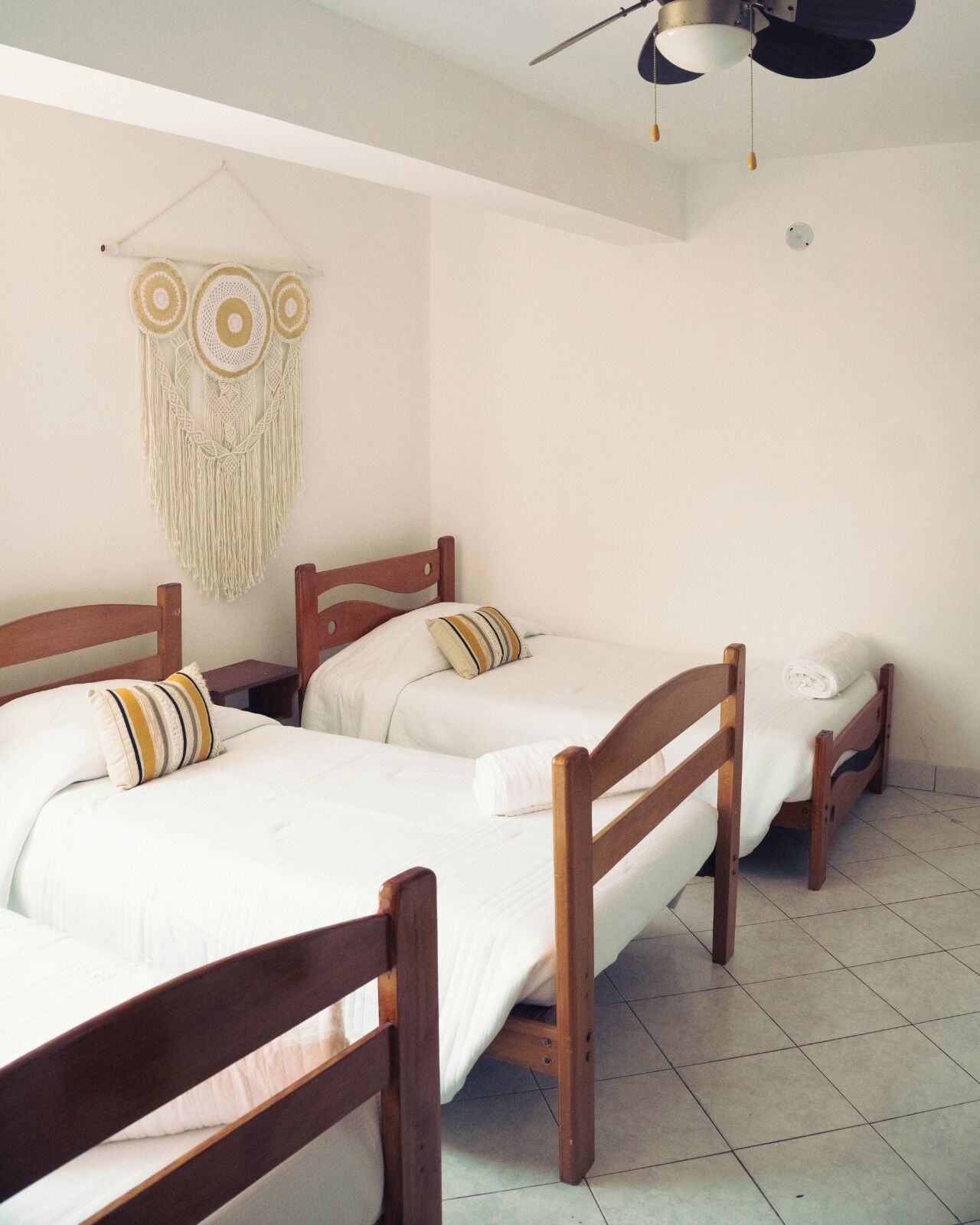 Una habitación con tres camas de madera, cada una con ropa blanca y almohadas a rayas. Se observa un tapiz de macramé sobre las camas.