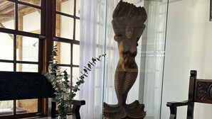Ein Holztisch mit einer Vase grüner Pflanzen, einer dekorativen Holzskulptur und Blick auf eine Bambuskonstruktion durch große Fenster.