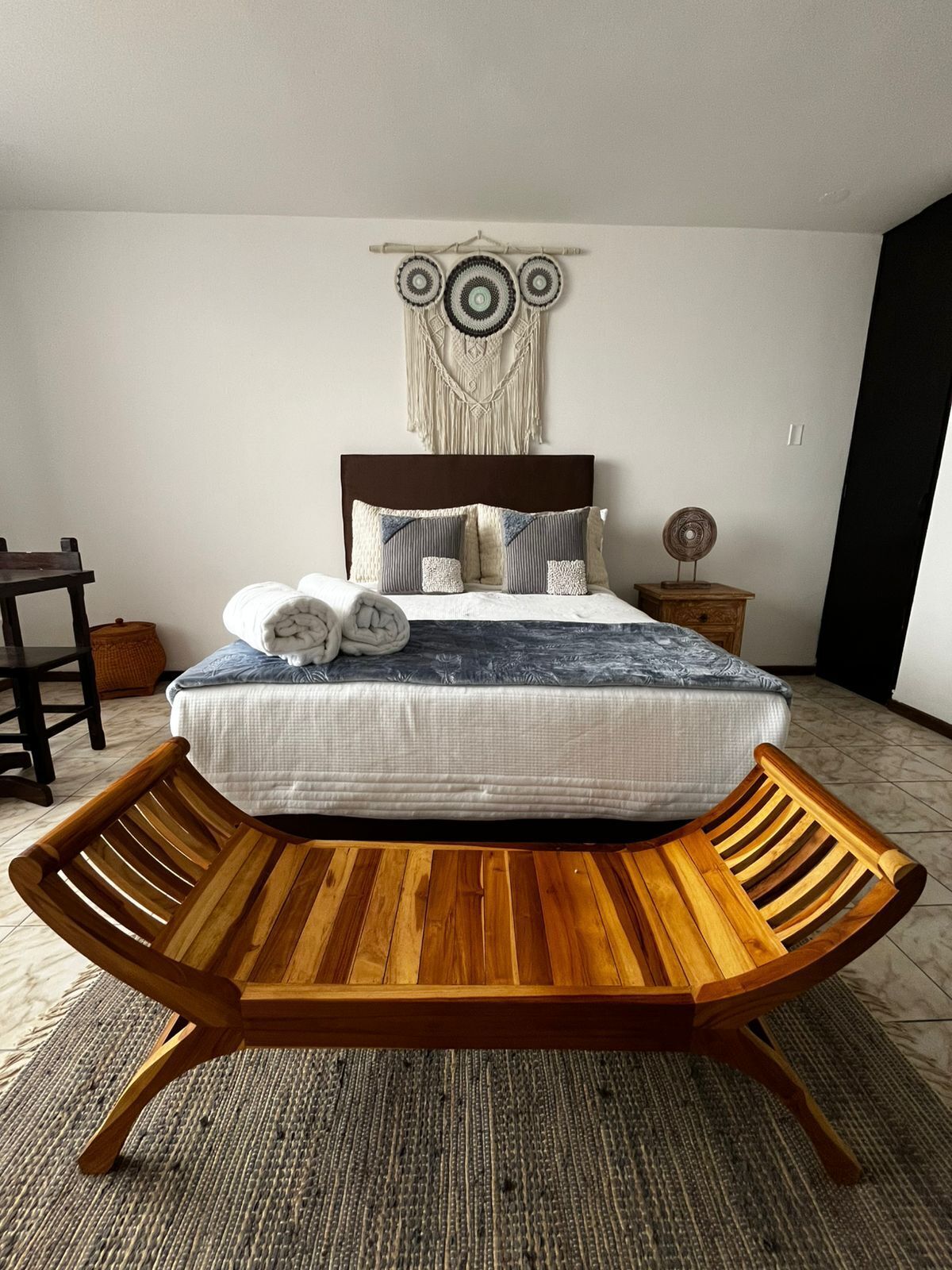 Un dormitorio con una cama, un banco de madera, un tapiz en la pared y una pequeña mesa de madera.