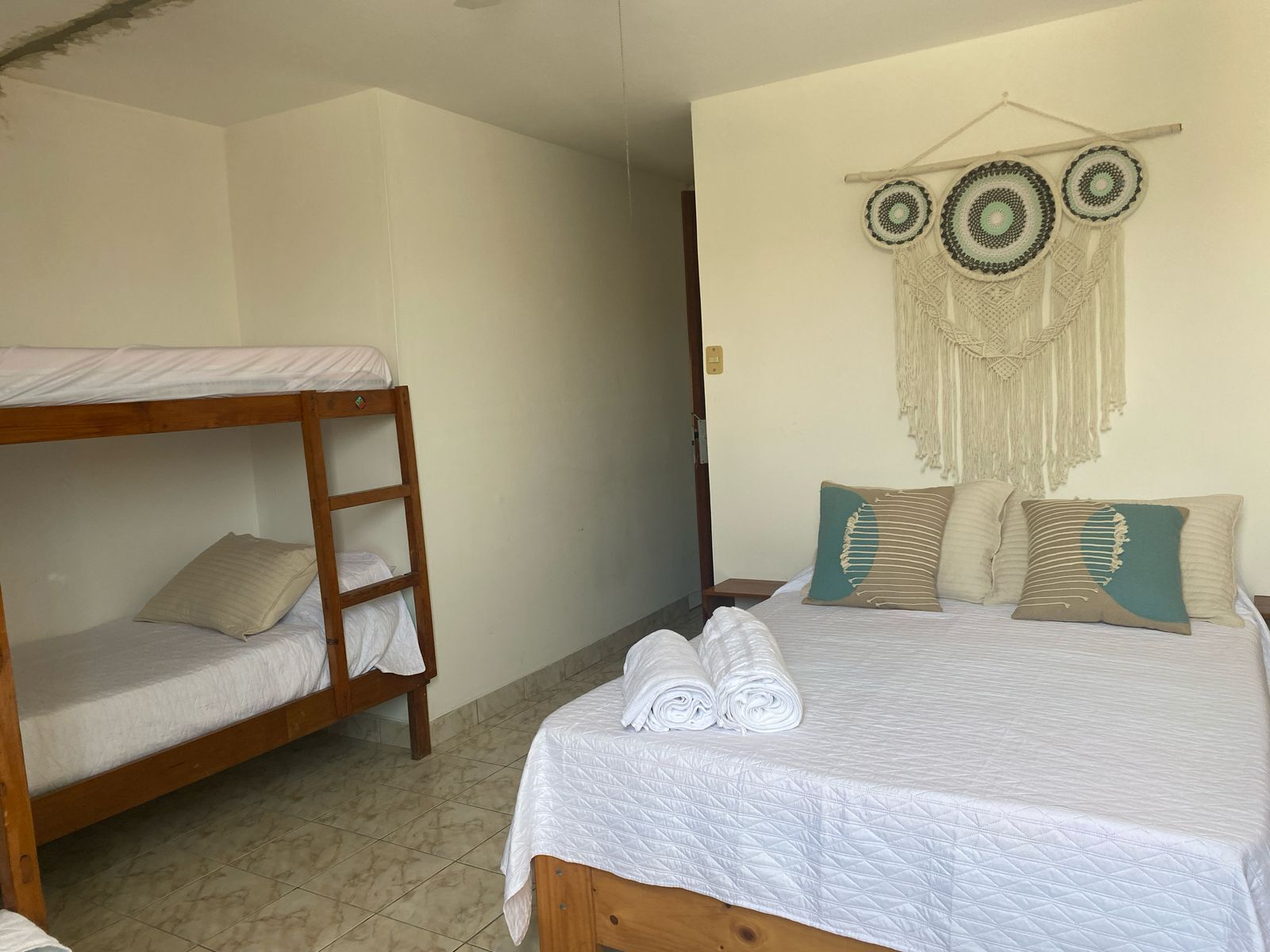 A room with a bunk bed, a single bed with rolled towels, and a wall with macrame decorations.