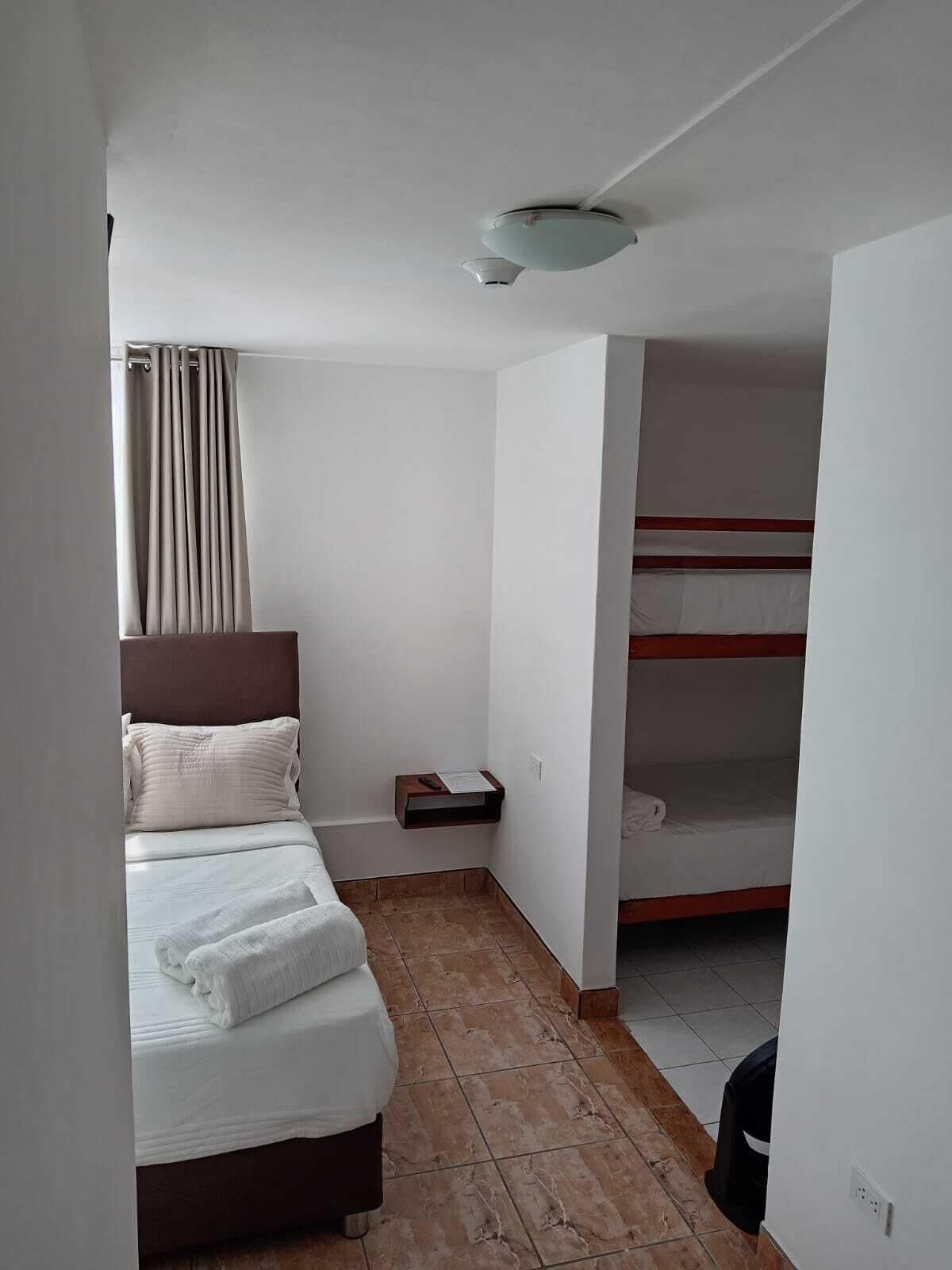 Habitación de hotel con cama, mesita de noche, estante pequeño, ventana con cortinas y una cama litera en la esquina.