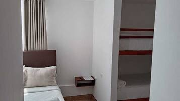 Habitación de hotel con cama, mesita de noche, estante pequeño, ventana con cortinas y una cama litera en la esquina.