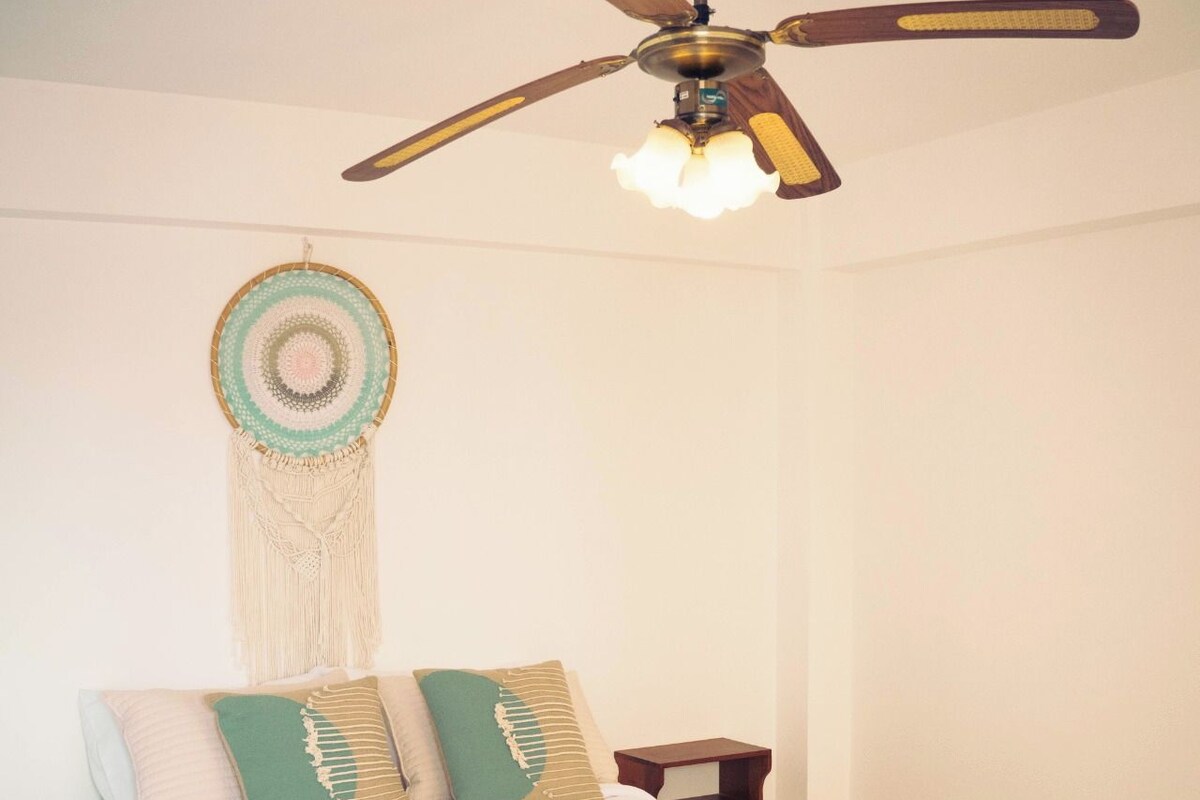 A bedroom with a bed, a ceiling fan, a wall hanging, and a nightstand.