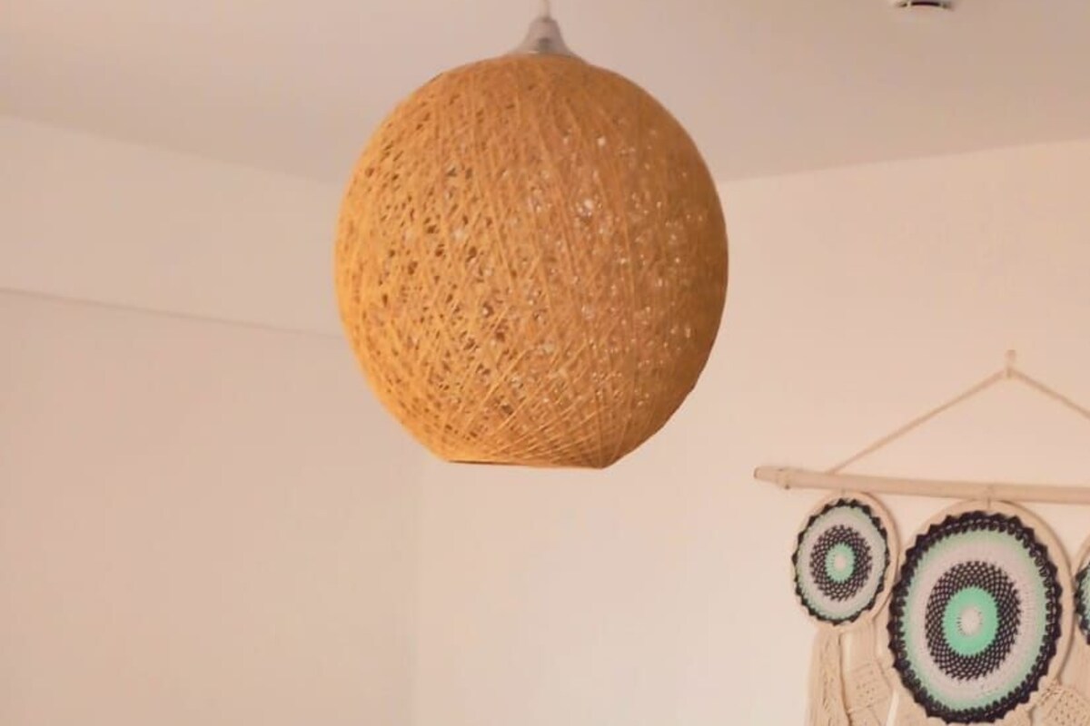 A neatly made bed with white linens and rolled towels, a woven hanging light fixture, and a wall hanging with circular patterns.